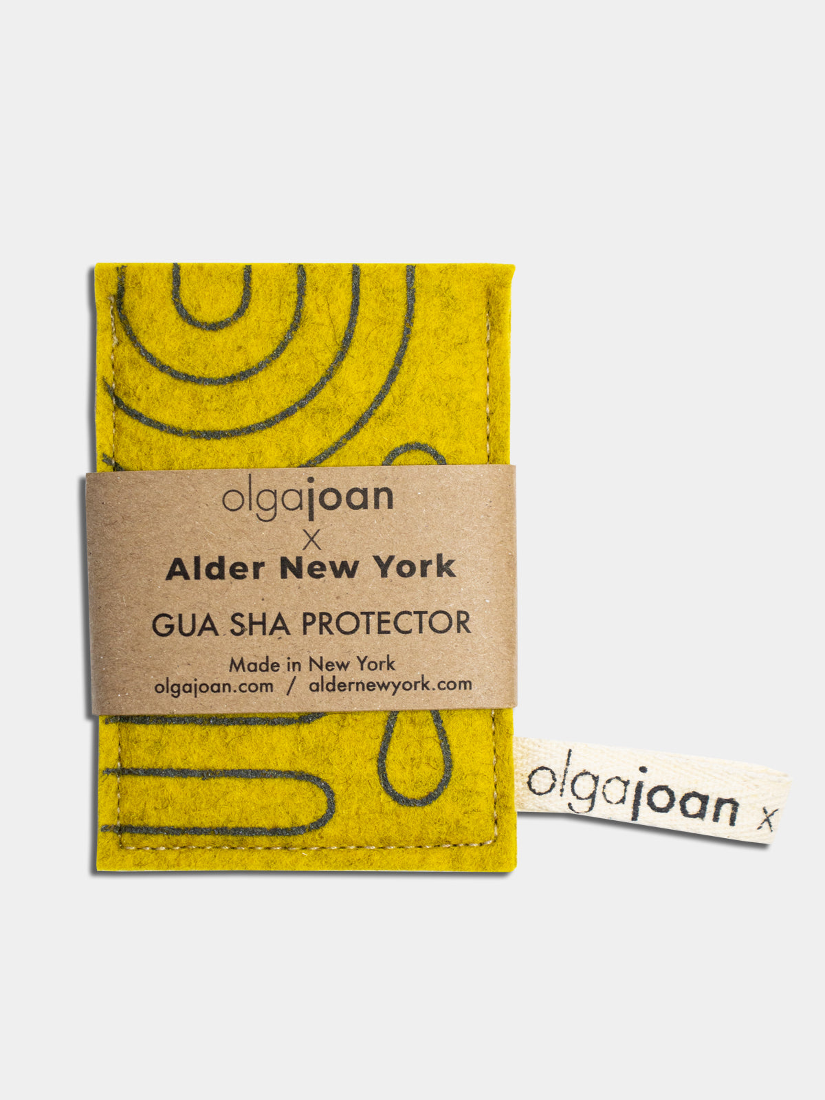 Yellow felted wool gua sha protector with a dark gray-green abstract pattern, shown with kraft paper packaging labeled “Olga Joan x Alder New York Gua Sha Protector — Made in New York.”